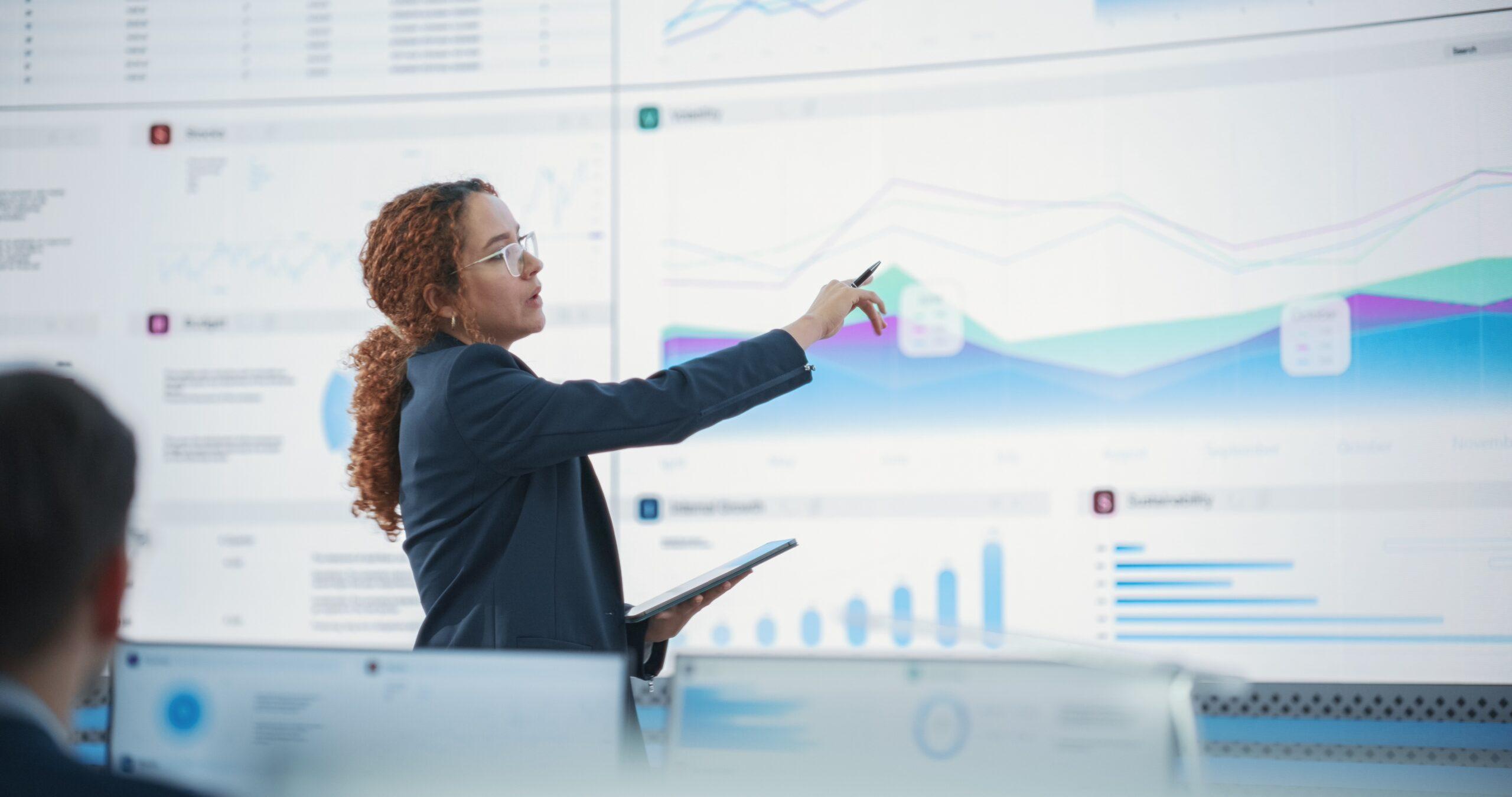 A woman in a blazer presents data visualizations on a wall in front of desktop computers