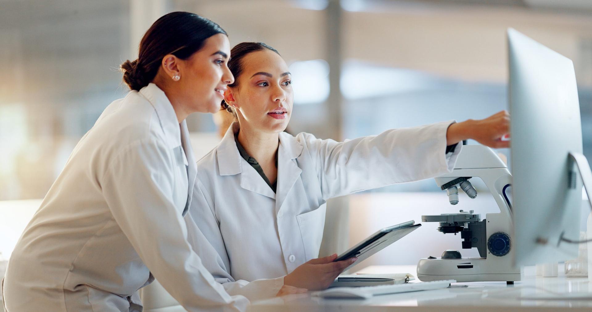 Two women in lab coats study a computer screen. One is pointing to something on the screen while the other looks on.