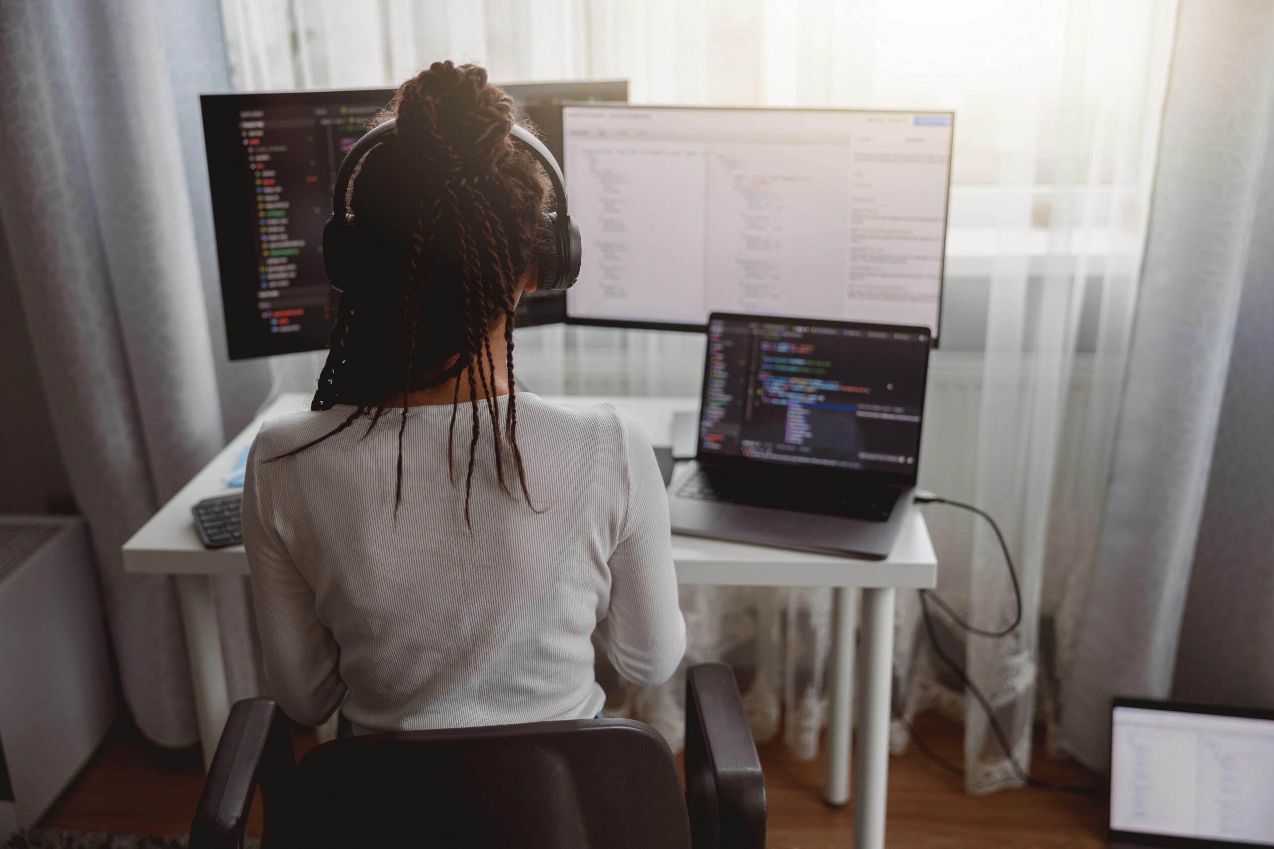 woman coder sitting at table coding on laptop looking at two computer monitors