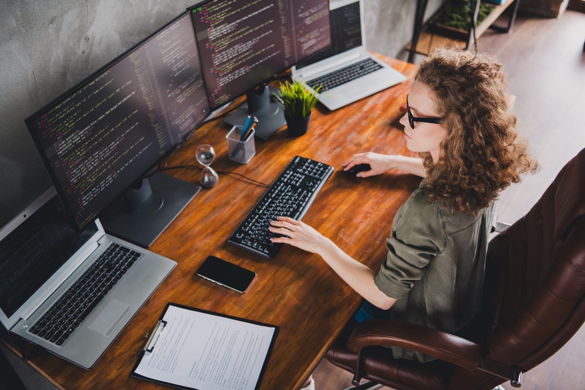 female software developer sitting at a workstation in front of several monitors with code